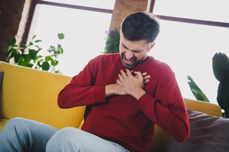 Young man in casual attire experiencing chest discomfort in modern apartment setting during daylightの写真素材