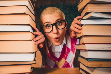 Young woman with short hair and glasses poses creatively between stacks of books, wearing a colorful...