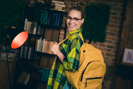 Charming young woman with blond hair and glasses smiling indoors wearing a checkered shirt and...