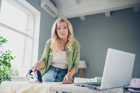 Mature woman with blond hair ironing clothes in the living room while engaging in housework activities, wearing casual outfit.の写真素材