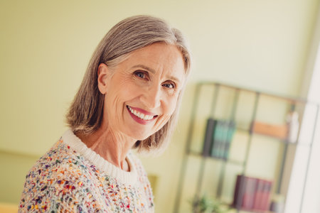 Charming mature woman in a stylish sweater smiling happily in a cozy living room. Embracing a joyful...