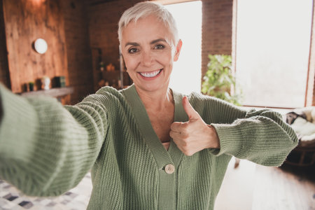 Charming elderly woman with short grey hair gives thumbs up and smiles warmly inside cozy living...
