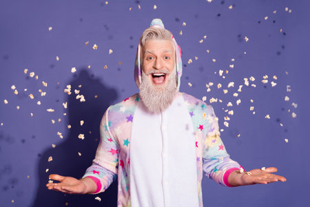Joyful man in colorful unicorn pajama surrounded by flying popcorn on purple background