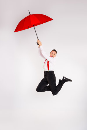 A young man joyfully jumps with a red umbrella, expressing happiness and energy in elegant fashion on a white backgroundの写真素材