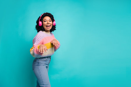 Excited young woman dancing in a colorful sweater with headphones on turquoise background
