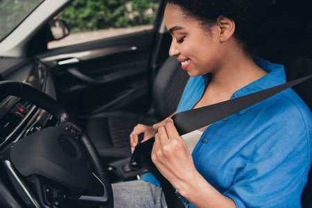 Young woman fastening seatbelt in car while smiling during a sunny day, symbolizing road safety and...