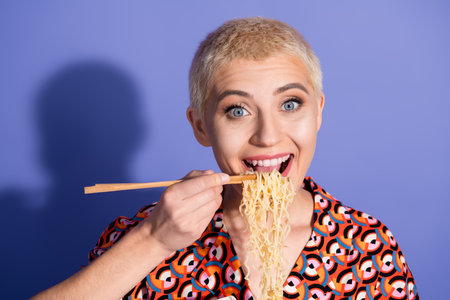 Young woman with a cheerful expression enjoys noodles with chopsticks against a purple backgroundの写真素材