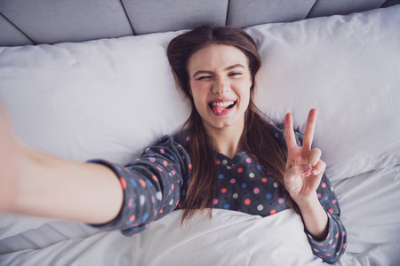 Young woman posing playfully in cozy pajamas while relaxing in a comfortable bed, expressing happiness and energyの写真素材