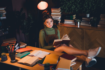 Young woman in a green turtleneck and brown jumper sits comfortably with a book in a cozy room filled with booksの写真素材
