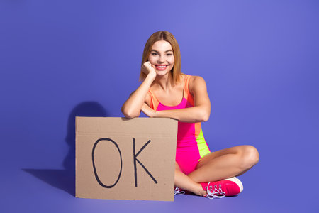 Smiling young woman wearing a vibrant sporty jumpsuit sitting against a purple background holding a cardboard sign saying OKの写真素材