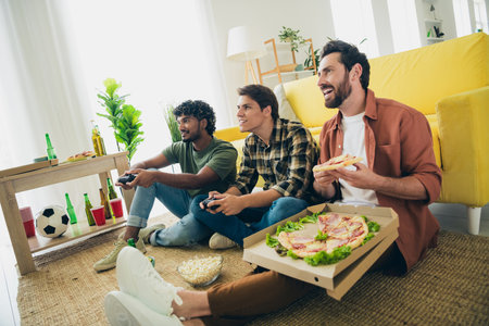 Photo of company three friends sit floor eat piza play videogame hang out weekend modern interior...