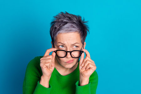 Thoughtful mature woman in green sweater with glasses against a vibrant blue background, showing a curious expressionの写真素材