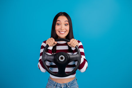 Smiling young woman holding a steering wheel, standing against a vibrant blue background, expressing...