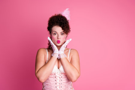 Young woman in vintage corset posing on pink background with surprised expression, blending historical fashion elementsの写真素材