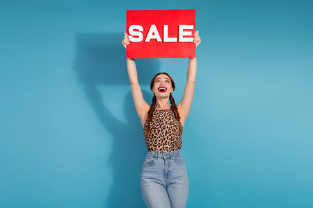 Cheerful young woman holding a red sign against a blue background, expressing excitement.の写真素材