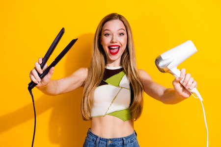 Cheerful young woman posing with hair styling tools on a bright yellow background, expressing fun, joyの写真素材