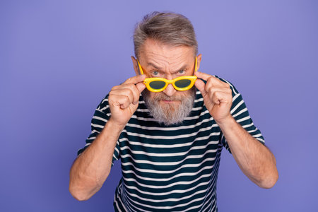 Stylish elderly man wearing yellow sunglasses and striped shirt posing against a violet background...