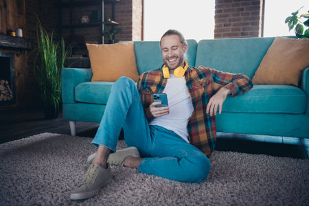 Casual young man relaxing on a sofa with a mobile phone and headphones in a stylish living room atmosphereの写真素材