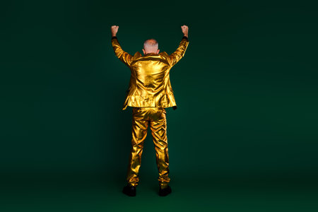 Elegant mature man celebrating in golden suit with arms raised against green backgroundの写真素材