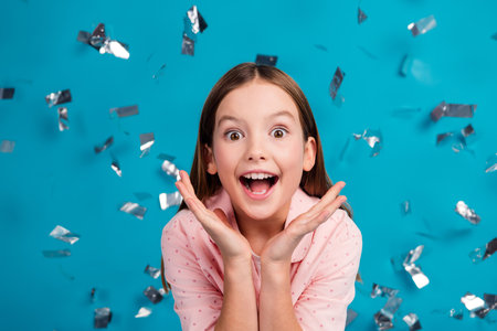 Excited young girl celebrating with a joyful face and confetti flying on a vibrant blue backgroundの写真素材