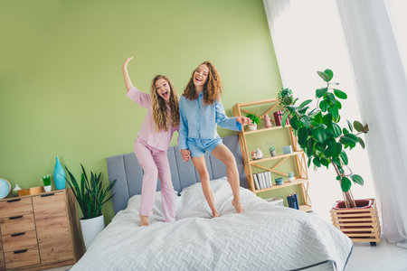 Full length photo of two pretty young females stand bed dancing dressed enjoy wake up modern cozy day light bedroom home interiorの写真素材
