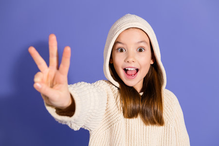 Charming young girl in a casual hoodie smiling joyfully against a purple background, holding up...