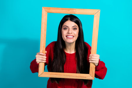 Attractive young woman in a red knit sweater holding an empty wooden frame against a vibrant...