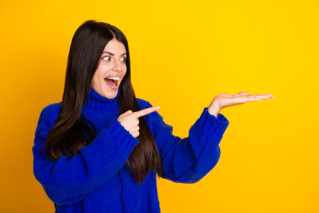 Smiling woman wearing blue sweater points to her hand against a vibrant yellow backgroundの写真素材