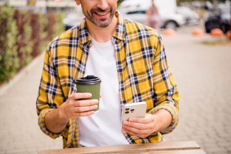 Young smiling man holding a coffee cup and smartphone outdoors wearing a yellow checkered shirt, enjoying leisure timeの写真素材