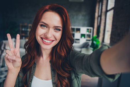 Photo of nice young ginger girl show v-sign take selfie photo wear khaki clothes enjoy modern cozy interior flat indoorsの写真素材