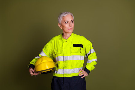 Mature woman in safety workwear holding a yellow hardhat against a khaki background, showcasing strong determinationの写真素材