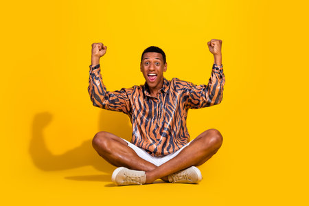 Excited young man in printed shirt sitting cross-legged and celebrating on bright yellow backgroundの写真素材