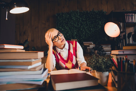 Young woman wearing stylish argyle waistcoat sits amidst stacks of books, peering through chic glassesの写真素材