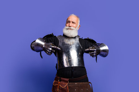 Elderly man dressed in medieval knight armor posing against a vibrant purple backgroundの写真素材