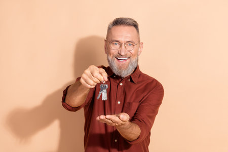 Smiling mature man holding keys and gesturing a hand invitation against a beige background, illustrating homeownership or real estateの写真素材