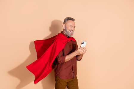 Confident mature man wearing a vibrant red cape while smiling and using his smartphone against a beige backgroundの写真素材