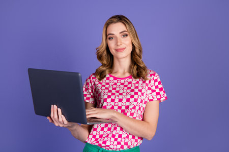 Young woman with a cheerful expression holding a laptop, standing against a vibrant purple background dressed in a trendy patterned outfit.の写真素材