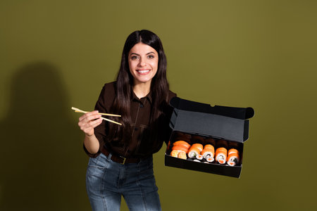 Attractive brunette woman holding sushi box with chopsticks and smiling against green backgroundの写真素材