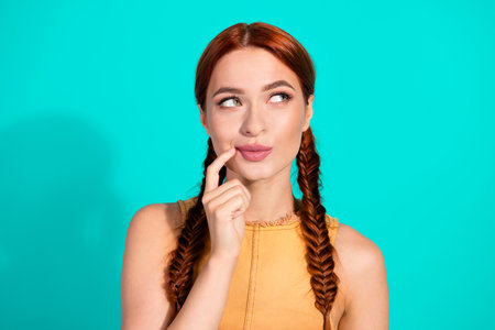 Portrait of a young woman with red braids in a yellow top posing against a teal background, displaying curiosity and playful expressionの写真素材