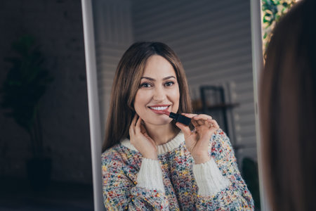 Young woman at home applying lipstick while smiling in front of a mirrorの写真素材