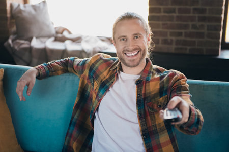 Smiling young man with beard relaxing at home on a comfortable sofa, wearing a stylish checkered...