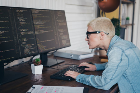 Young woman coding in a home office setup wearing a denim shirt and focused on multi-screen...