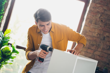 Young man assembling furniture indoors with a smile, showcasing stylish casual attire in a modern home environmentの写真素材