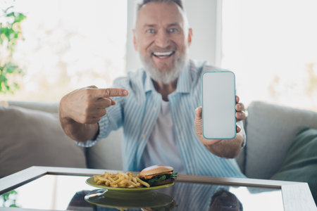 Smiling elderly man relaxing indoors with a plate of food, showcasing smartphone screen, enjoying lifestyle and daylight in a cozy home.の写真素材