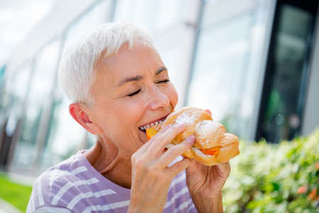 Elderly woman enjoying a delicious pastry outdoors on a sunny day in the city, with a happy and carefree expression on her lovely face.の写真素材