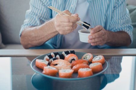 Elderly man enjoying sushi with chopsticks at home in a casual setting during daytimeの写真素材