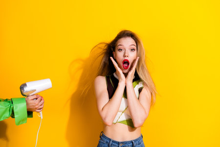 Young woman expressing surprise as her hair is blown with a hairdryer against a bright yellow backgroundの写真素材
