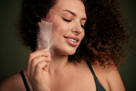 Young woman with natural curly hair holding a feather, radiating confidence and grace, embracing her unretouched beautyの写真素材