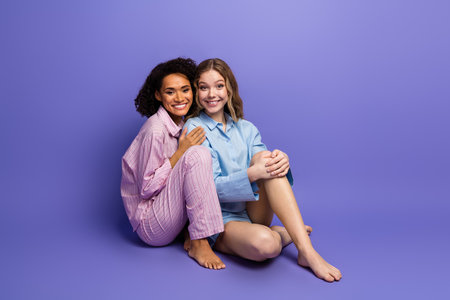 Two young women in pajamas sitting together on a purple background, smiling and enjoying a cozy atmosphereの写真素材