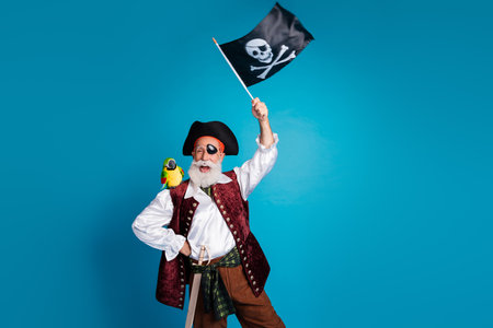 Photo portrait of senior funny pirate grandpa wave flag wear masquerade party costume isolated blue color backgroundの写真素材